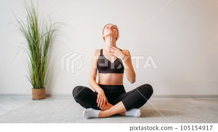 Morning exercise. Home fitness. Indoor training. Relaxed inspired athletic girl stretching neck muscles sitting cross-legged on floor at light studio interior copy space background. Morning exercise. Home fitness. Indoor training. Relaxed inspired athletic girl stretching neck muscles sitting cross-legged on floor at light studio interior copy space background. 105154917