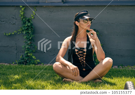 Woman posing sitting on a green grass of a recreation center or hotel with a swimming pool. Dressed in a black swimsuit, sun visor and sunglasses. Woman posing sitting on a green grass of a recreation center or hotel with a swimming pool. Dressed in a black swimsuit, sun visor and sunglasses. 105155377