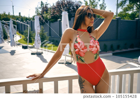 Portrait of a woman having rest in a recreation center or hotel with a swimming pool. Dressed in a colorful swimsuit and sunglasses. Portrait of a woman having rest in a recreation center or hotel with a swimming pool. Dressed in a colorful swimsuit and sunglasses. 105155428