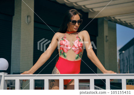 Portrait of a woman having rest in a recreation center or hotel with a swimming pool. Dressed in a colorful swimsuit and sunglasses. 105155431