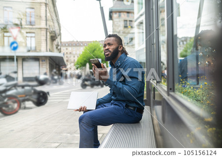 Bearded young african american man sitting on the bench with a phone in hands Bearded young african american man sitting on the bench with a phone in hands 105156124