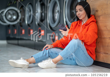 Young woman listening music with smartphone and earphones waiting in laundry room. Young woman listening music with smartphone and earphones waiting in laundry room. 105156219