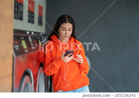 Young woman listening music with smartphone and earphones waiting in laundry room. Young woman listening music with smartphone and earphones waiting in laundry room. 105156243