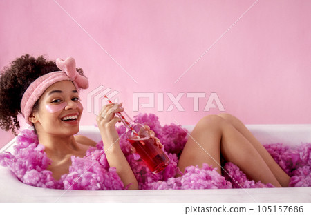 Attractive young female african with terry headband and patches under her eyes is sitting in bath on pink background 105157686