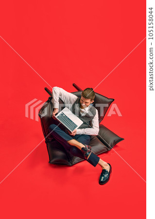 Businessman, employee sitting on armchair in weird, uncomfortable position, working on laptop against red studio background 105158044