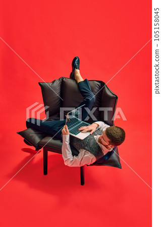 Businessman, employee sitting on armchair in weird, uncomfortable position, working on laptop against red studio background 105158045