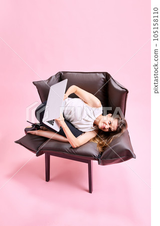 Businesswoman lying in uncomfortable, weird posign on armchair, feeling irritated, working on laptop against pink studio background Businesswoman lying in uncomfortable, weird posign on armchair, feeling irritated, working on laptop against pink studio background 105158110