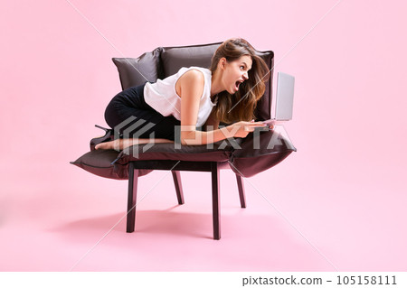 Project manager, employee sitting in weird pose on armchair, looking on laptop and shouting with anger against pink studio background 105158111