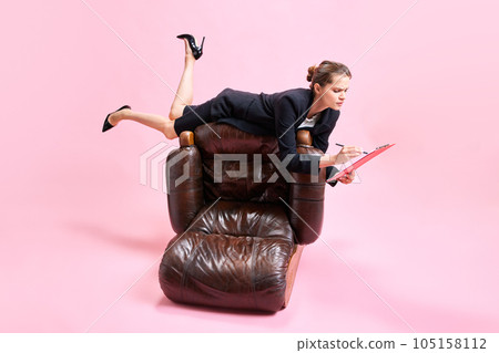 Business woman lying on top of armchair and looking through documents against pink studio background Business woman lying on top of armchair and looking through documents against pink studio background 105158112