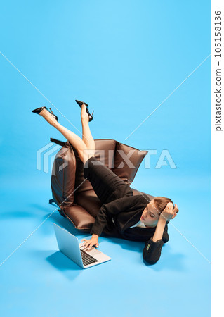 Young woman, employee lying on fallen armchair and working on laptop against blue studio background. Weird position Young woman, employee lying on fallen armchair and working on laptop against blue studio background. Weird position 105158136