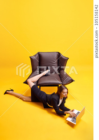 Young business woman, employee lying on floor near chair and working online on laptop against bright yellow background Young business woman, employee lying on floor near chair and working online on laptop against bright yellow background 105158142