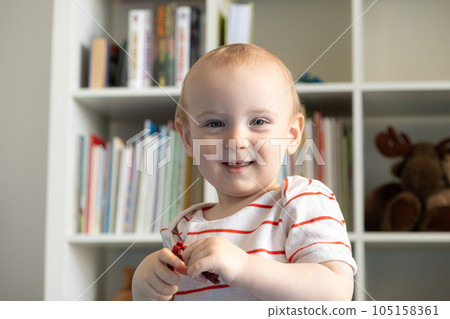 Portrait of a one-year-old toddler with a toy in his hands in a children's room, smiling, looking at the camera. 105158361