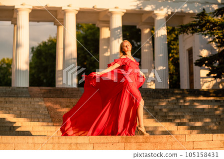 A woman in a long red dress against the backdrop of sunrise, bright golden light of the sun's rays. The concept of femininity, harmony. 105158431