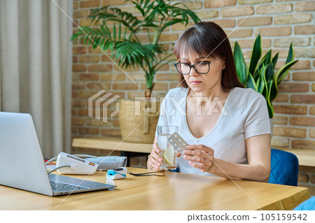 Mature woman with pills and glass of water at table at home 105159542