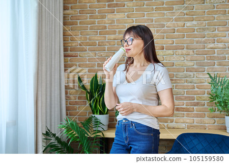 Middle aged woman drinking fermented milk product from bottle, near window 105159580