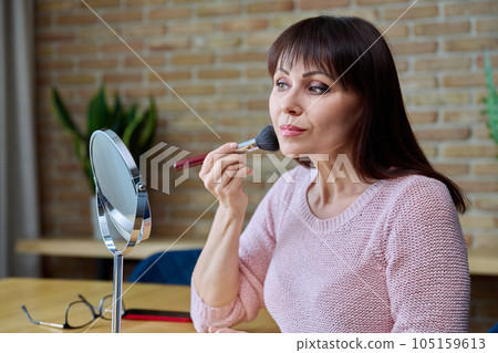 Middle-aged woman with make-up mirror cosmetics doing makeup at home 105159613