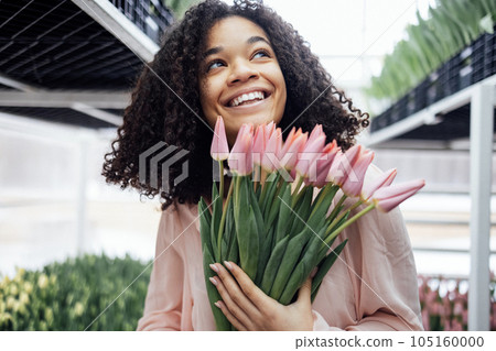 Beautiful romantic smiling african ethnicity girl holding bouquet of pink tulips in greenhouse. Close up of feminine young afro woman among rows and shelves of fresh flowers in winter garden. Beautiful romantic smiling african ethnicity girl holding bouquet of pink tulips in greenhouse. Close up of feminine young afro woman among rows and shelves of fresh flowers in winter garden. 105160000