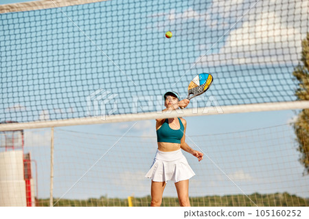 Dynamic image of young woman playing beach tennis, hitting ball with racket. Outdoor training on warm summer day 105160252