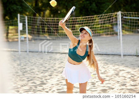 Young gsportive girl in cap and sportswear playing beach tennis, hitting ball with racket. Outdoor training on warm summer day Young gsportive girl in cap and sportswear playing beach tennis, hitting ball with racket. Outdoor training on warm summer day 105160254