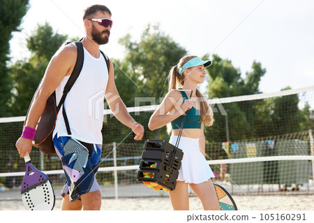 Young man and woman going to play tennis on beach on warm summer day. Sportive young people in sportswear carrying tennis equipment 105160291