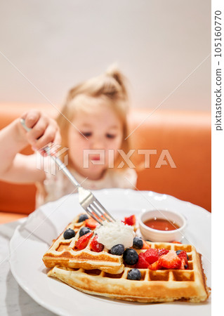 Selective focus in the foreground. Happy 3 year old girl eat. Breakfast in cafe. Classic Viennese waffles with ice cream, berries and Maple syrup. Table in the restaurant. 105160770