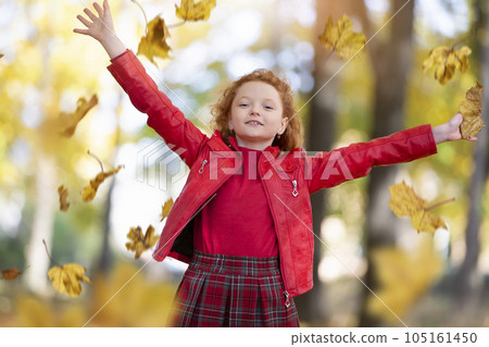 Beautiful red-haired girl in the autumn park scatters maple leaves. 105161450