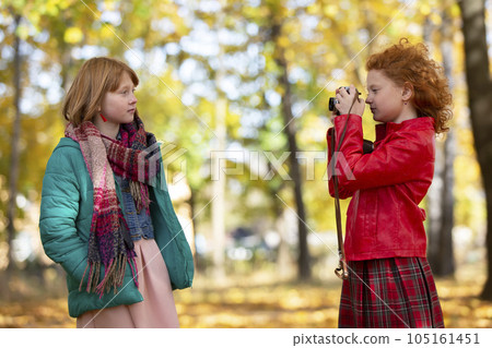 Two red-haired girls girlfriends photograph each other in the autumn park with a retro camera. 105161451