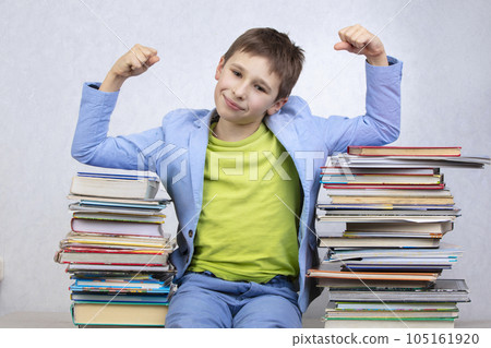 Happy smart school boy sits between stacks of books and shows a sign of strength. Overcoming learning difficulties. High school. Happy smart school boy sits between stacks of books and shows a sign of strength. Overcoming learning difficulties. High school. 105161920