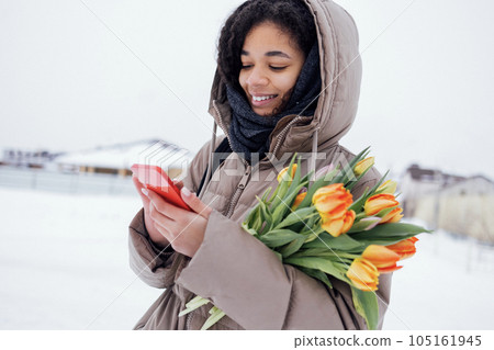 Young enchanting african woman in warm casual clothes holds phone and bouquet of orange tulips. Young enchanting african woman in warm casual clothes holds phone and bouquet of orange tulips. 105161945