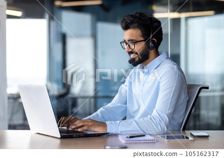 A young Indian businessman is working in the office on a laptop wearing a headset. A young Indian businessman is working in the office on a laptop wearing a headset. 105162317