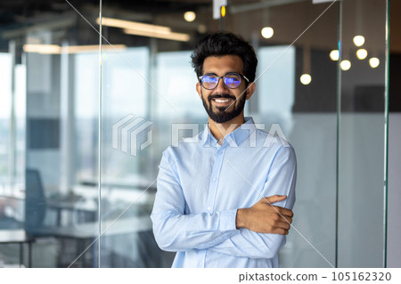Portrait of a young Indian male designer, engineer, architect who is wearing glasses and a blue shirt smiling standing in the office and looking at the camera. Portrait of a young Indian male designer, engineer, architect who is wearing glasses and a blue shirt smiling standing in the office and looking at the camera. 105162320