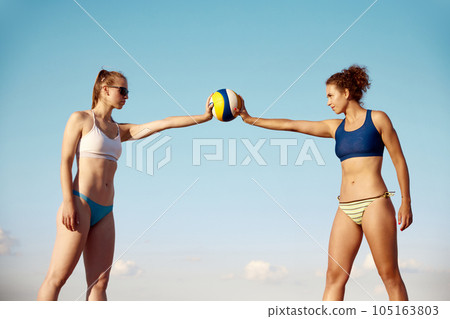 Two sportive young womsn, with fit, slim body in swimsuit holding volleyball ball over blue sky background 105163803