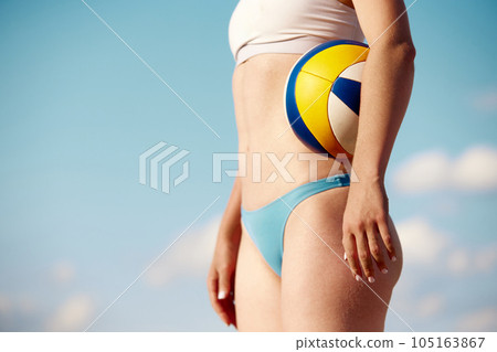 Cropped image of female slim, fit body in swimsuit. Model standing with volleyball ball over blue sky background Cropped image of female slim, fit body in swimsuit. Model standing with volleyball ball over blue sky background 105163867