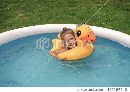 A cute child smiling looks at the camera while swimming on a circle in the shape of a duck in a small inflatable rubber swimming pool in the backyard A cute child smiling looks at the camera while swimming on a circle in the shape of a duck in a small inflatable rubber swimming pool in the backyard 105167540