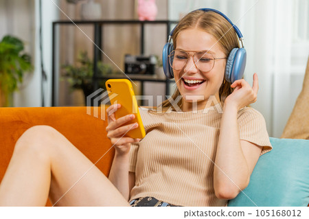 Happy young woman in wireless headphones relaxing lying on sofa at home listening favorite music 105168012