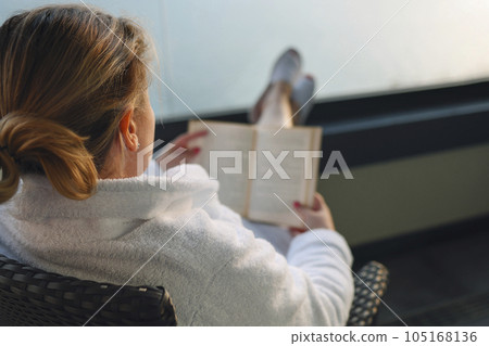 Close up of portrait of young woman in white coat sitting in wicker chair on balcony and reading book. 105168136
