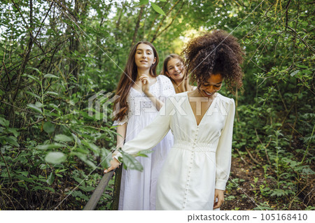 Three happy pretty smiling teenage girls in white summer elegant dresses walk along path in forest or park. Three happy pretty smiling teenage girls in white summer elegant dresses walk along path in forest or park. 105168410