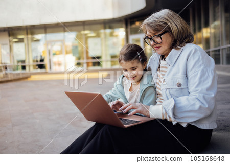 A middle-aged woman and a little girl are sitting outdoors with a laptop. 105168648