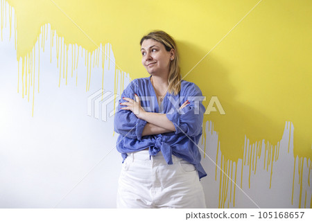 A young blonde woman in casual clothes stands at a white wall unpainted with yellow paint in a room of her house. 105168657