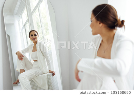 A beautiful girl with evening make-up in white clothes sits on an ottoman and smiles. Reflection of a young woman and panoramic windows in the mirror. 105168690