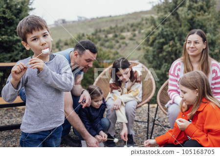 Big happy family roasting mashmallows outdoors. Little cute boy eating hot marshmallow on stick. Big happy family roasting mashmallows outdoors. Little cute boy eating hot marshmallow on stick. 105168705