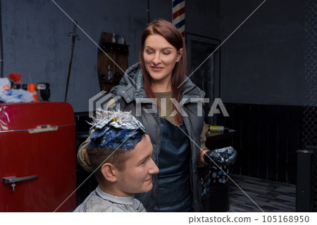 Satisfied beautiful brunette adult girl European professional barbershop worker in the process of dyeing hair in color for a young guy client 105168950