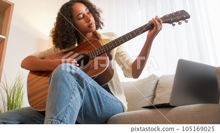 Guitar hobby. Music practice. Weekend leisure. Focused woman playing chords on acoustic string instrument sitting sofa at home room. 105169025