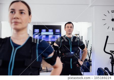 Couple of people in ems suits are preparing for workout in fitness club or gym. Young man and woman stand with their hands on their belts. 105169224