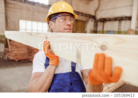 Good-looking male builder carrying wooden plank at construction site Good-looking male builder carrying wooden plank at construction site 105169413