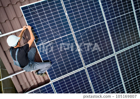 Man technician installing photovoltaic solar moduls on roof of house. Electrician in helmet building solar panel system outdoors. Concept of alternative and renewable energy. Aerial view. Man technician installing photovoltaic solar moduls on roof of house. Electrician in helmet building solar panel system outdoors. Concept of alternative and renewable energy. Aerial view. 105169807