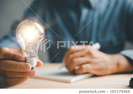 Businessman captures a good idea, holding a light bulb in his left hand. Symbol of solution and success. Working in an inspiring office, the author finds inspiration with a pen on paper. 105170468