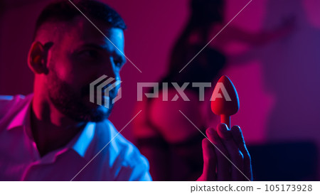 Bearded caucasian man holding an anal plug. Close-up of female buttocks in underwear. Sexual preferences of the couple. 105173928