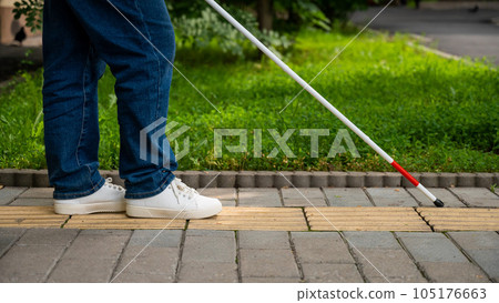 A blind woman walks outdoors using a cane along a tactile yellow tile. A blind woman walks outdoors using a cane along a tactile yellow tile. 105176663