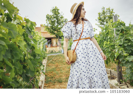 Young enchanting woman in summer sundress with floral pattern, hat and straw bag looks dreamily at sky. Young enchanting woman in summer sundress with floral pattern, hat and straw bag looks dreamily at sky. 105183060